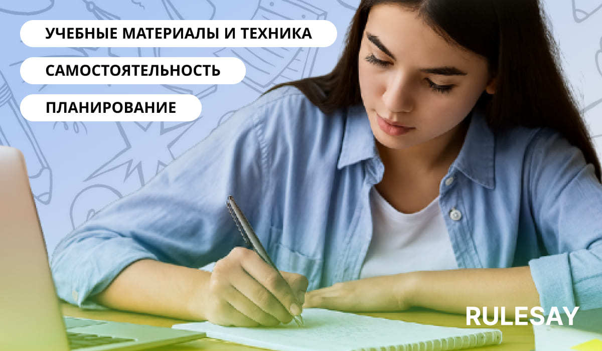 Молодая девушка в голубой рубашке сосредоточенно делает записи в тетради за столом с ноутбуком